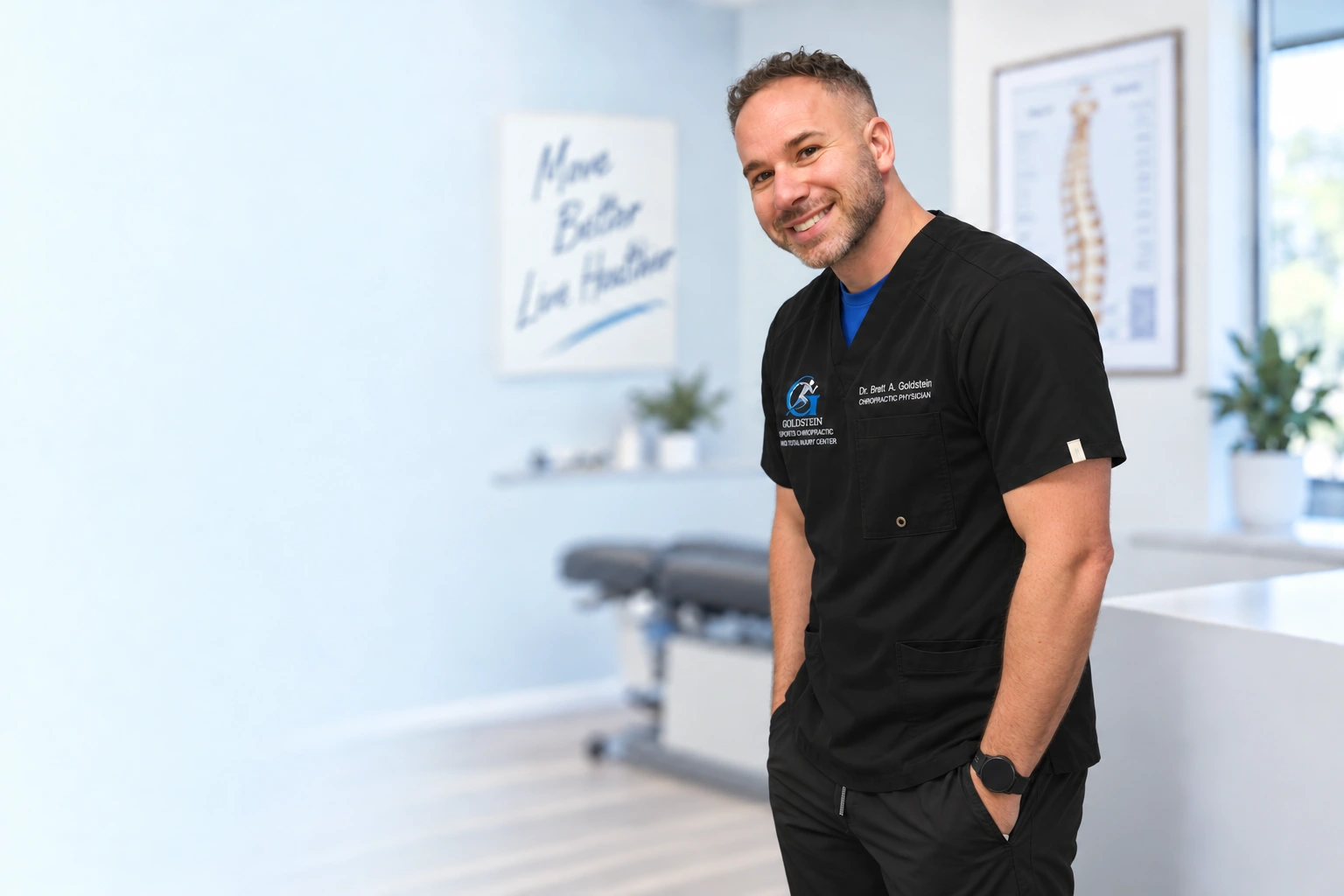 Dr. Brett Goldstein smiling in black branded scrubs and a blue undershirt, with hands resting in his pockets