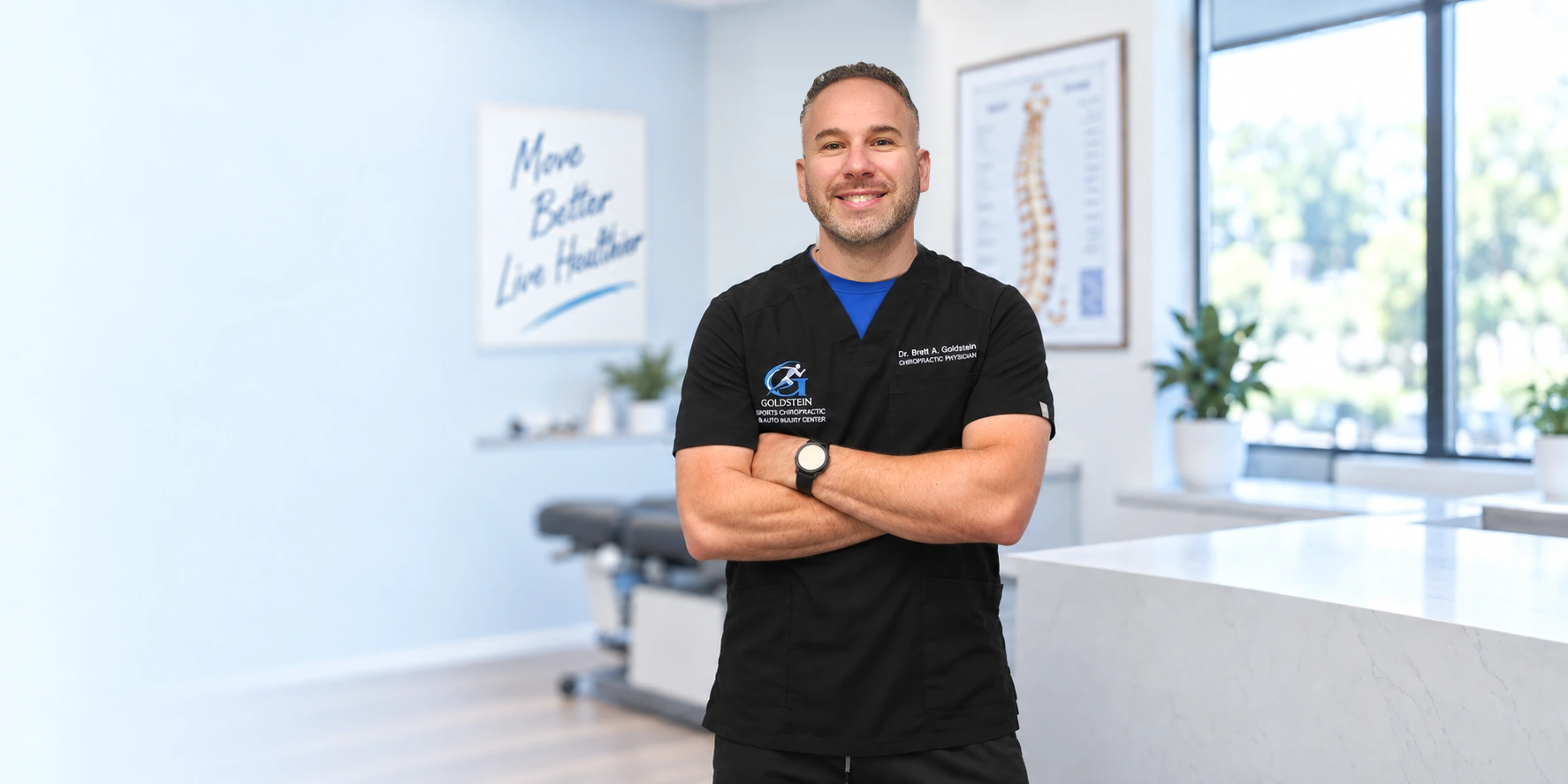 Dr. Brett A. Goldstein smiling with arms crossed, wearing black scrubs embroidered with the Goldstein Sports Chiropractic and Auto Injury Center logo